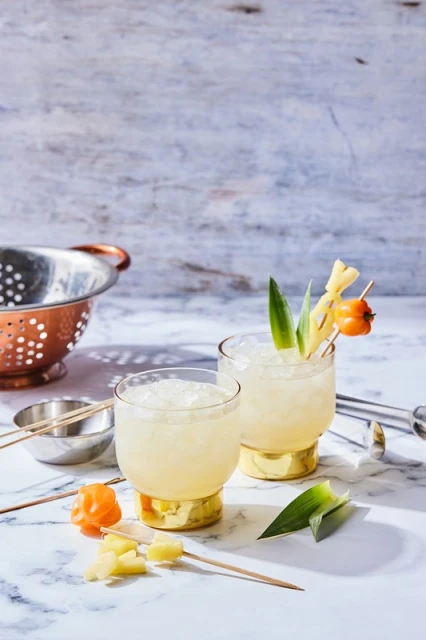 Two tropical cocktails garnished with pineapple and peppers sit on a marble surface, with a strainer and bar tools adding to the scene.