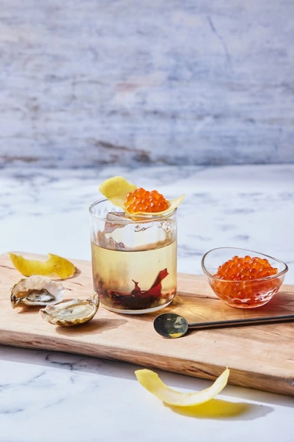 Caviar and oysters arranged on wooden board. Includes a glass with drink, spoon, lemon peel, bowl of caviar. White background.