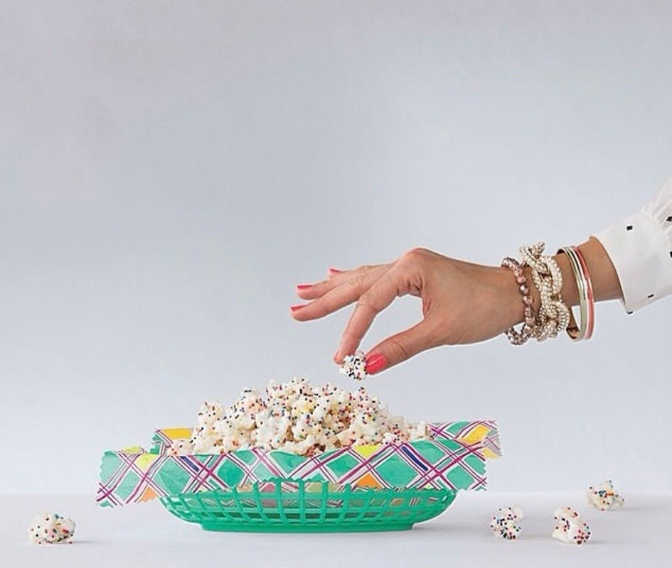 Woman's hand holding a piece of sprinkle popcorn over a green basket filled with popcorn on a white table in a white room.