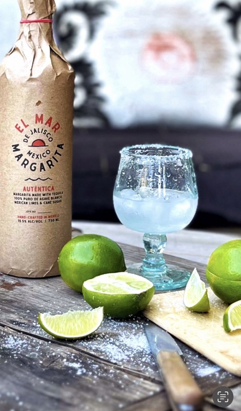 Bottle of El Mar Margarita tequila next to salt-rimmed glass and limes on rustic wooden surface, ideal for cocktail or beverage ad.