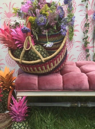 Woven basket filled with fresh purple flowers and pineapples resting on a pink velvet bench outdoors.