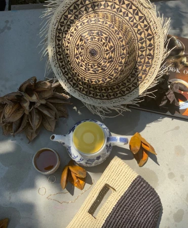 Top-down prop styling shot featuring a woven straw hat, a blue and white patterned teapot, a woven clutch, and dry seed pods.