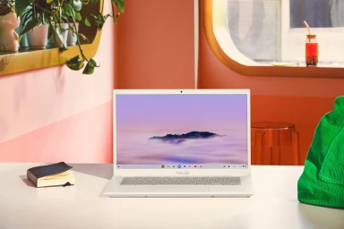 A white laptop centered on a white desk with a green bag, a small notebook, and pink and peach-toned walls in the background.
