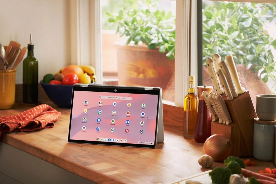 Silver laptop displaying app icons set on a wooden kitchen counter with a knife block, oil bottles, and a fruit bowl.