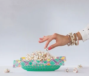 Hand reaching into a green plastic basket filled with popcorn covered in colorful sprinkles on white.