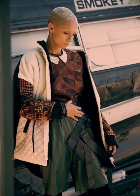Adult female in a conceptual fashion outfit poses outdoors with a neutral facial expression against an industrial, aged exterior backdrop.