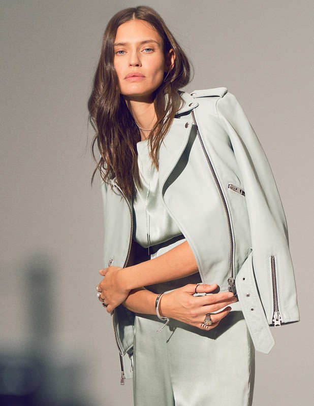Fashion shot of a brunette woman in a light green satin outfit, wearing a light green leather jacket, posing in a photo studio.