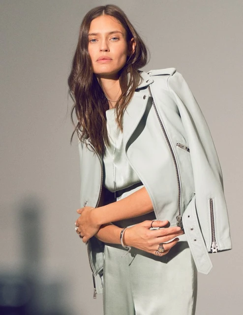 Fashion shot of a brunette woman in a light green satin outfit, wearing a light green leather jacket, posing in a photo studio.