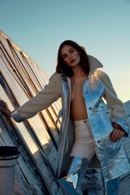 A woman in an avant-garde fashion coat and metallic boots poses on a rooftop with mirrored walls under a bright blue sky.