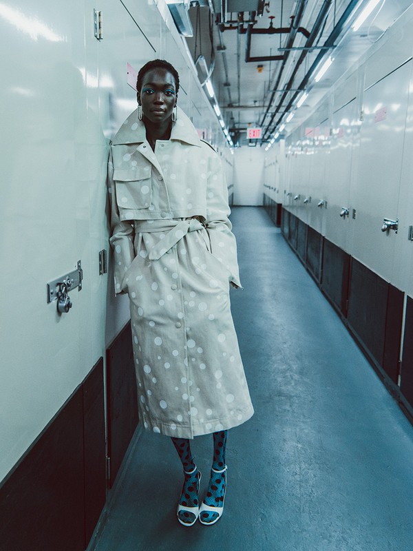 A tall, slim African-American model poses indoors against a white wall, wearing a beige polka-dot trench coat, patterned tights, and high heels.