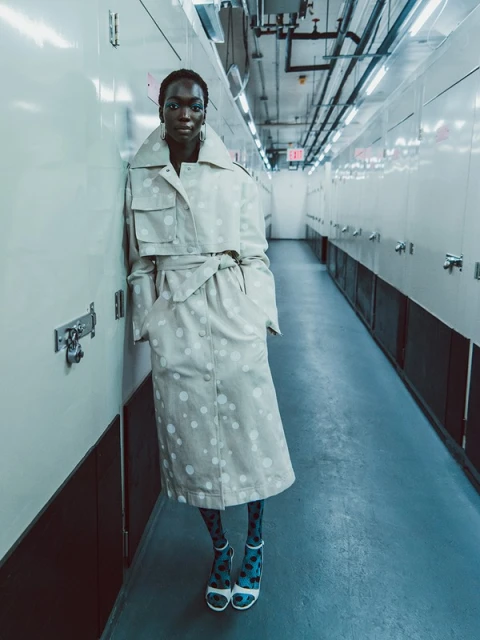 A tall, slim African-American model poses indoors against a white wall, wearing a beige polka-dot trench coat, patterned tights, and high heels.