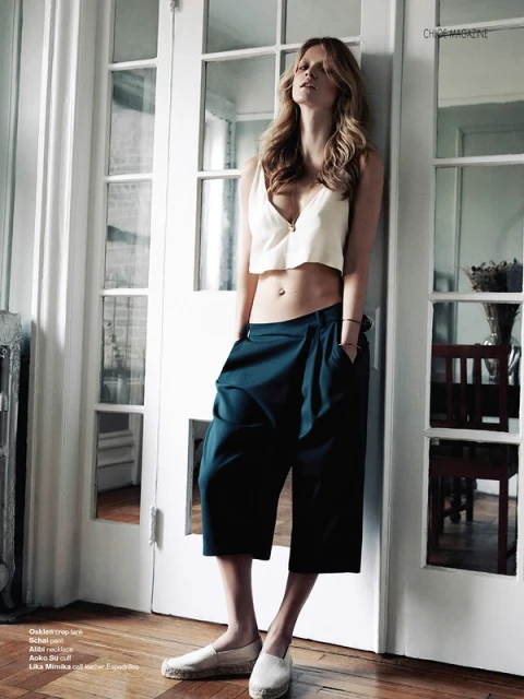 Indoor fashion shot of a woman in a cream top and blue culottes, leaning against a white door, with a wood floor visible.