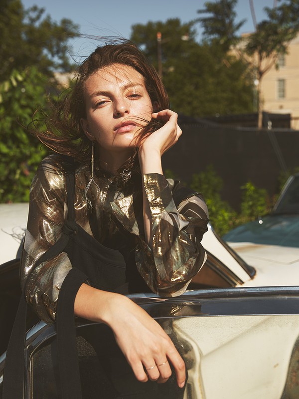 Woman with wind-blown hair, wearing a patterned top and resting against a classic car in an outdoor urban setting.