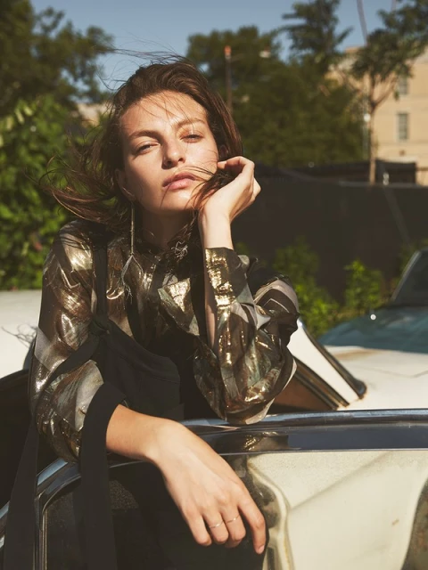 Woman with wind-blown hair, wearing a patterned top and resting against a classic car in an outdoor urban setting.