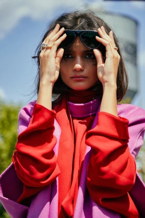 Close-up portrait of a woman adjusting dark sunglasses, wearing a vibrant red turtleneck and purple coat.