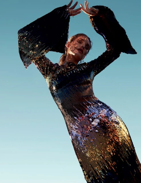 A woman in a long-sleeved, sequined dress stands against a clear blue sky, her hands raised above her head in a dynamic pose.