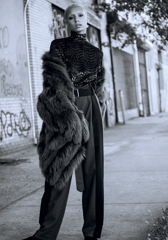 Fashionable woman poses in an urban setting. She wears a fur coat, reptile-print turtleneck, wide-leg trousers with a belt, and black footwear.