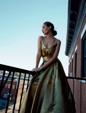 Woman in gold gown on balcony overlooking city buildings, bright and airy fashion shot with buildings and light blue sky background.