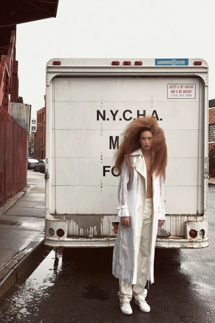 Adult female in a white outfit and large hair poses in front of a white truck on a city street with brick buildings.