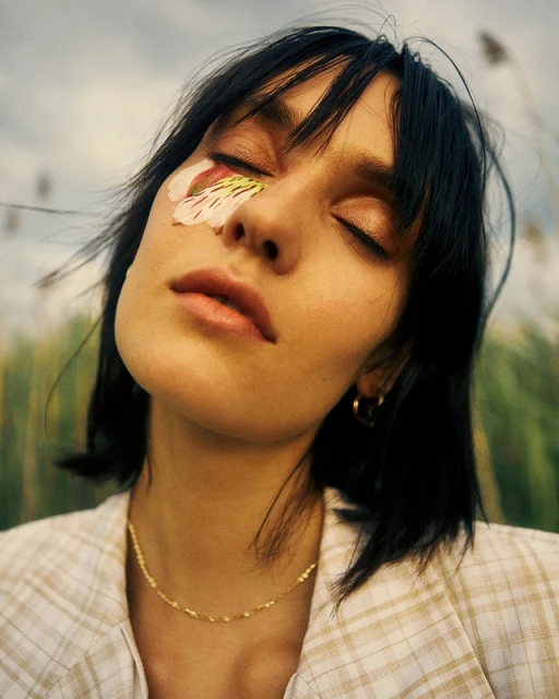 Close-up of a woman with black hair and closed eyes, wearing a tan blazer and gold necklace, with a flower on her face.