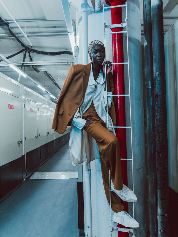 Stylish African woman in a brown suit and headscarf, posing on a metal ladder in an industrial space. Fashion editorial photography.