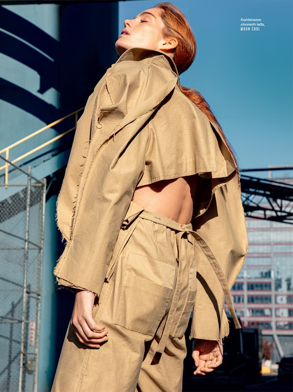 Model with reddish-brown hair wearing a khaki trench coat and trousers set outdoors under a clear blue sky.