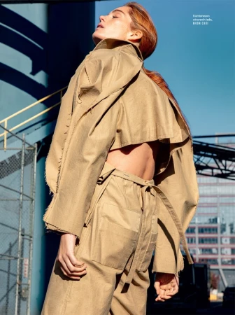 Model with reddish-brown hair wearing a khaki trench coat and trousers set outdoors under a clear blue sky.