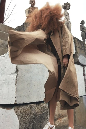 Model with voluminous red hair wearing a tan trench coat and white sneakers standing against a concrete background.