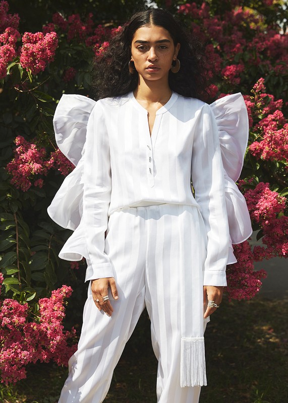 A woman in a white striped blouse and pants poses outdoors with pink flowers in the background, styled with a large statement sleeve.