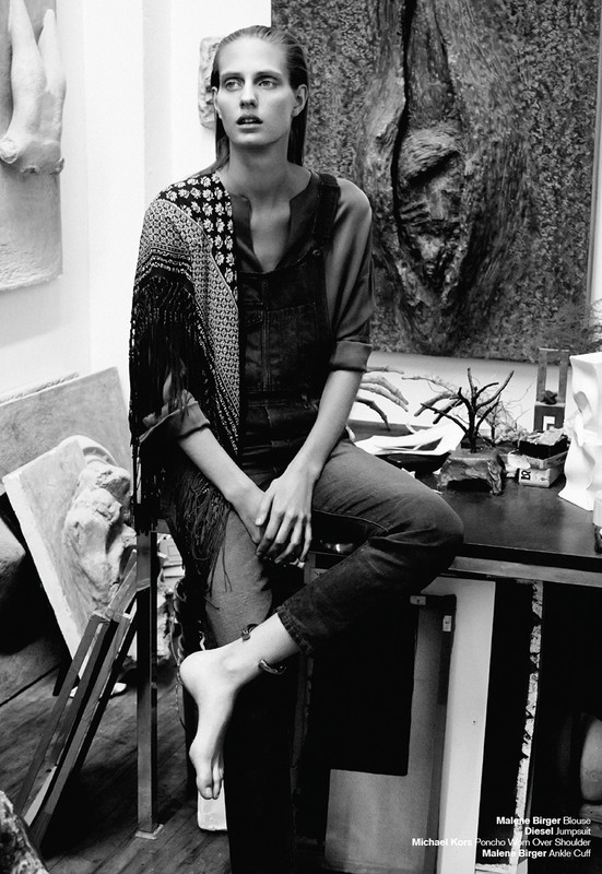 Black and white portrait of a bare-footed woman in overalls sitting on a table, with art sculptures and objects in the background.