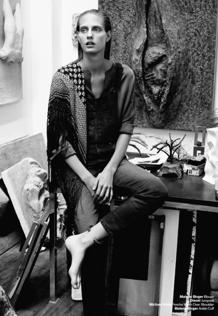 Black and white portrait of a bare-footed woman in overalls sitting on a table, with art sculptures and objects in the background.
