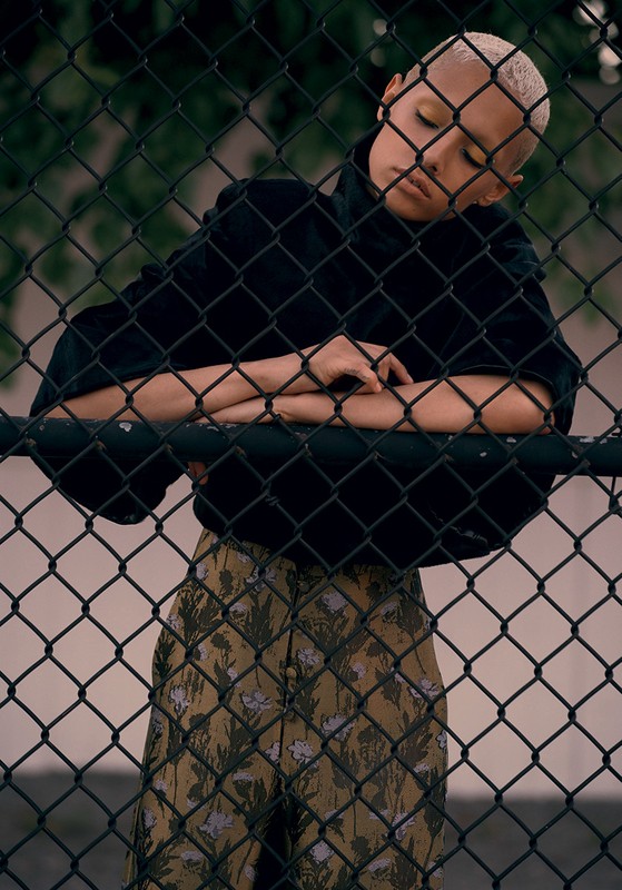 Portrait of a young adult with short blonde hair in a black jacket and patterned pants, leaning against a chain-link fence outdoors.