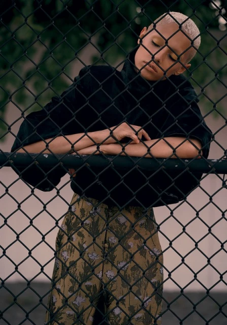 Portrait of a young adult with short blonde hair in a black jacket and patterned pants, leaning against a chain-link fence outdoors.