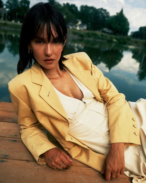 Stylish woman in a yellow jacket and cream dress poses on a wooden dock by a lake with trees in the background on a sunny day.