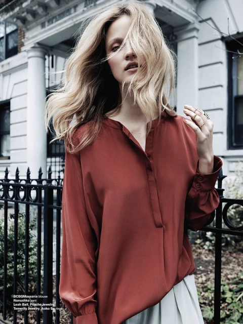 A stylish adult woman with wavy blonde hair poses outdoors in a rust-colored long-sleeved blouse with pleated skirt and rings.