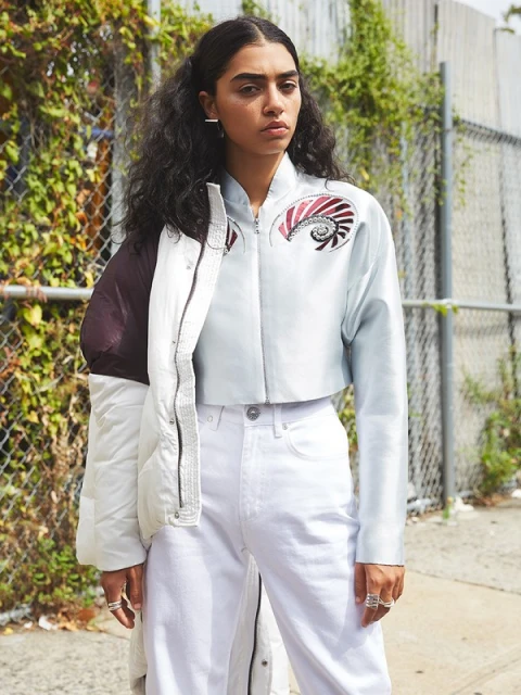 Stylish woman in light-blue jacket and white jeans poses against an urban fence with greenery in an eye-level outdoor shot.