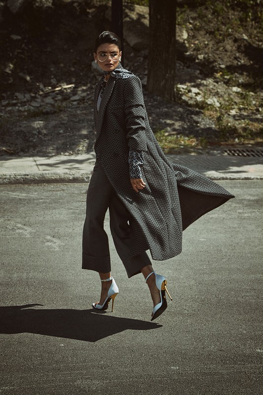 An adult female model in a grey coat and pants walks on a city street on a sunny day, wearing heels and glasses in a fashion editorial.