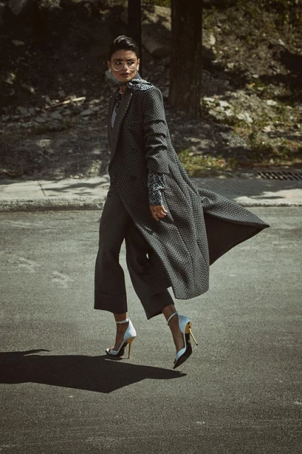 An adult female model in a grey coat and pants walks on a city street on a sunny day, wearing heels and glasses in a fashion editorial.