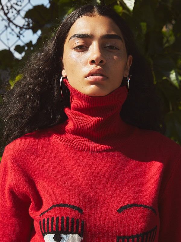 Close-up portrait of a woman with dark curly hair wearing a red turtleneck sweater with a graphic eye design, standing outdoors.