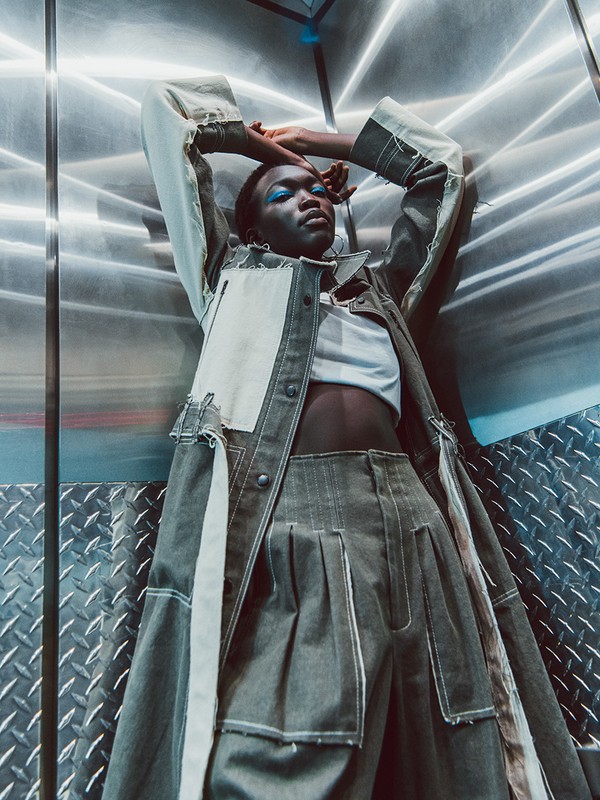 Fashion-forward, a young woman poses in modern denim outfit, lying in an industrial elevator with metallic walls and abstract lighting.