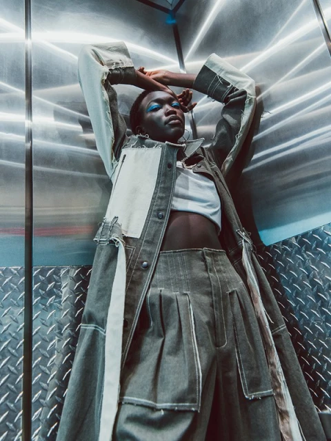 Fashion-forward, a young woman poses in modern denim outfit, lying in an industrial elevator with metallic walls and abstract lighting.