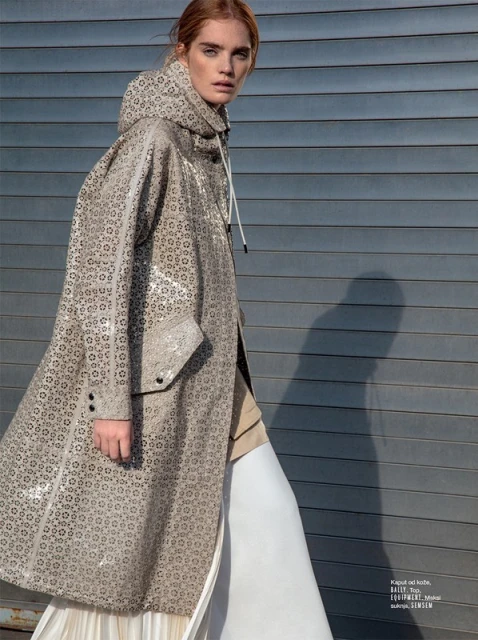 Adult woman fashion model poses in a patterned hooded coat, a long white dress, walking in an urban setting with a corrugated metal wall.