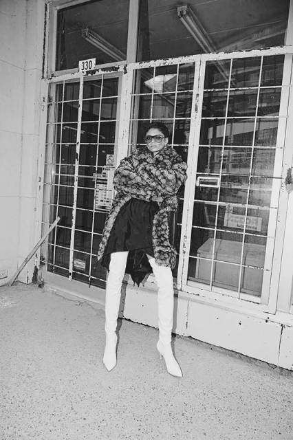 A woman in sunglasses, a patterned coat, and thigh-high boots poses by a barred window with a '330' sign in black and white.