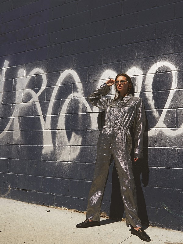 Woman in silver jumpsuit poses against graffiti-covered brick wall, wearing sunglasses, with shadows and bright sunlight.