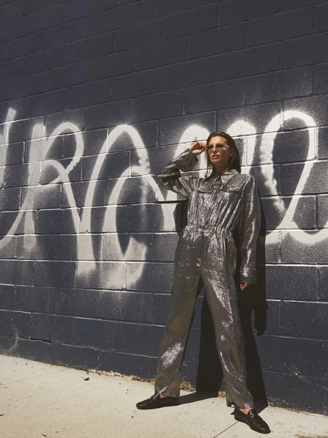 Woman in silver jumpsuit poses against graffiti-covered brick wall, wearing sunglasses, with shadows and bright sunlight.