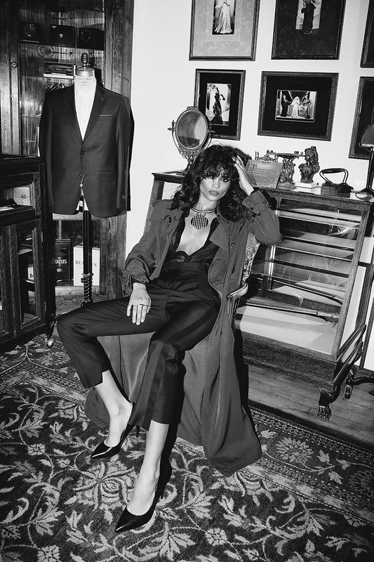 A woman poses in a chair in a dark, monochrome, vintage-style indoor scene, wearing a satin jumpsuit and dark shoes with a deep-V neckline.