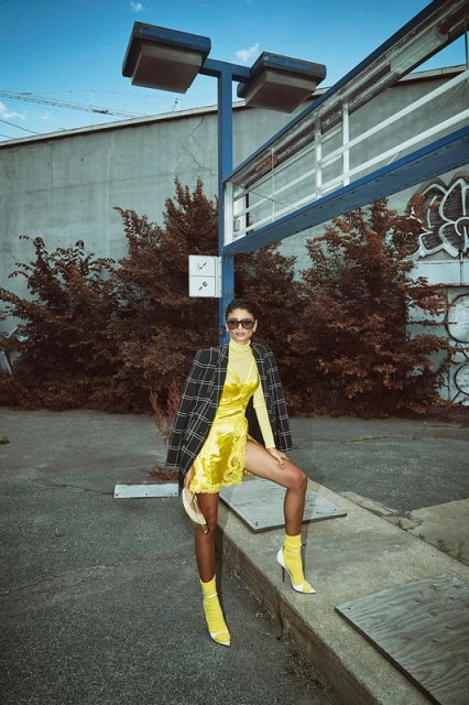 An adult female model poses in a yellow dress and boots in an outdoor urban setting, with a plaid jacket and a blue industrial structure.