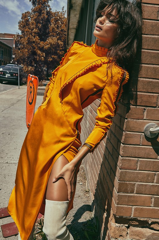 A woman in an orange dress and white boots leans against a brick wall on a sunny street with trees and a black car in the background.
