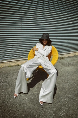 Model wearing a white suit and black bucket hat seated in a bright yellow woven chair outdoors in Los Angeles