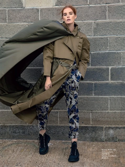 Fashion editorial shot of a red-haired woman posing in a tan trench coat and floral pants against a grey brick wall, urban fashion.